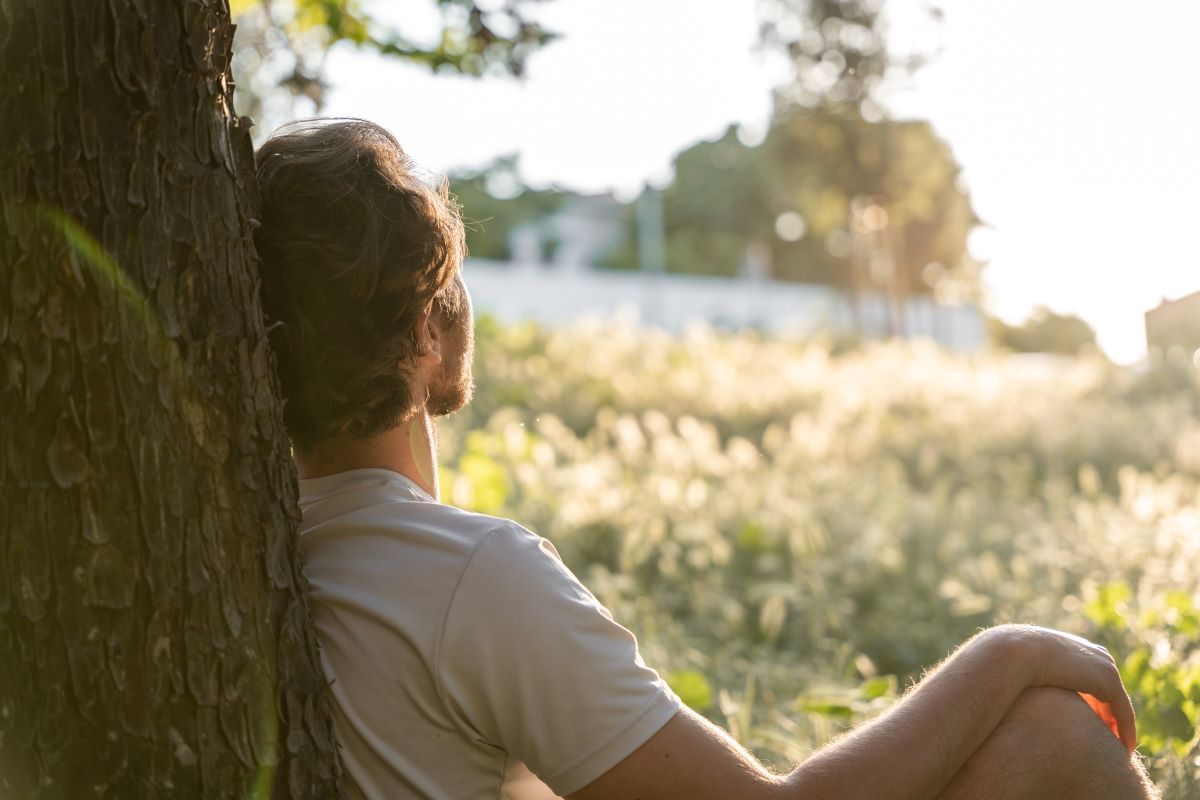A relaxed young man sits against a tree in nature.