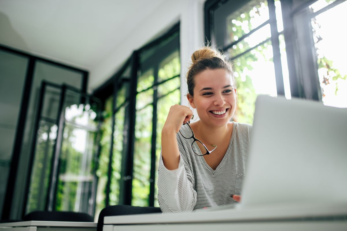 A smiling woman holding glasses looks at her laptop.