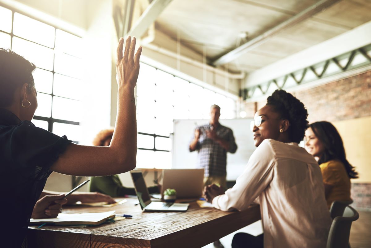 Professionals in a meeting; a woman raises her hand.