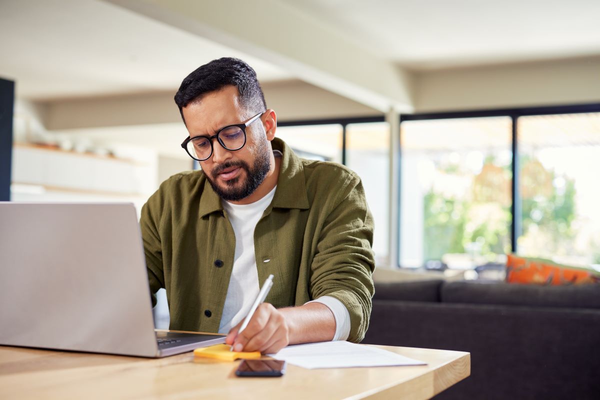 A man sits at a laptop and writes on a notepad.