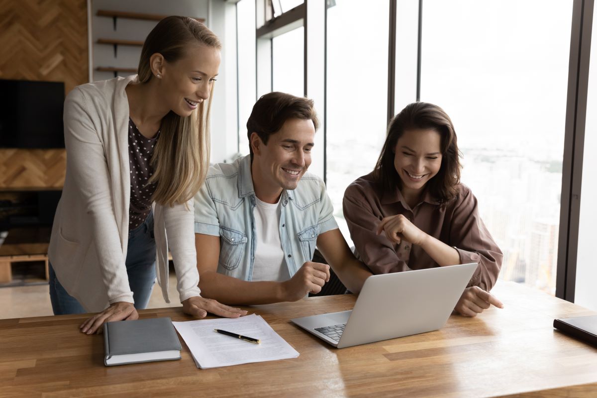 Three smiling work colleagues look at a laptop.