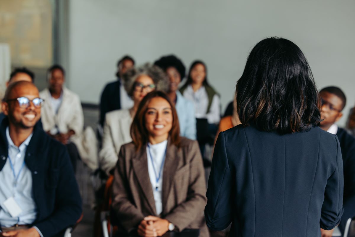 A group of smiling professionals listen to a woman presenting.
