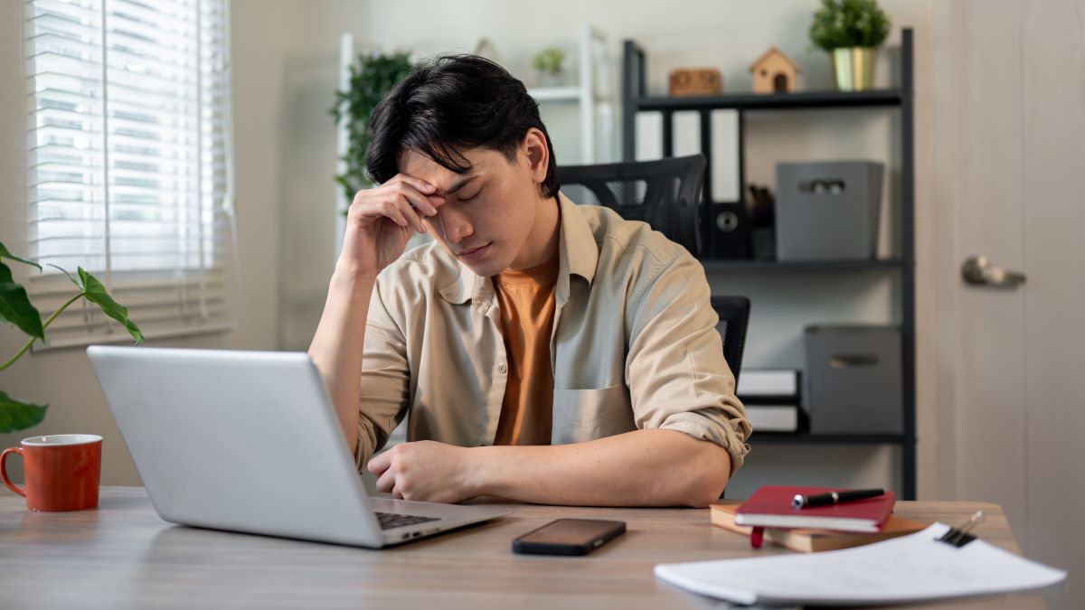 A young man with a headache sits at a laptop.
