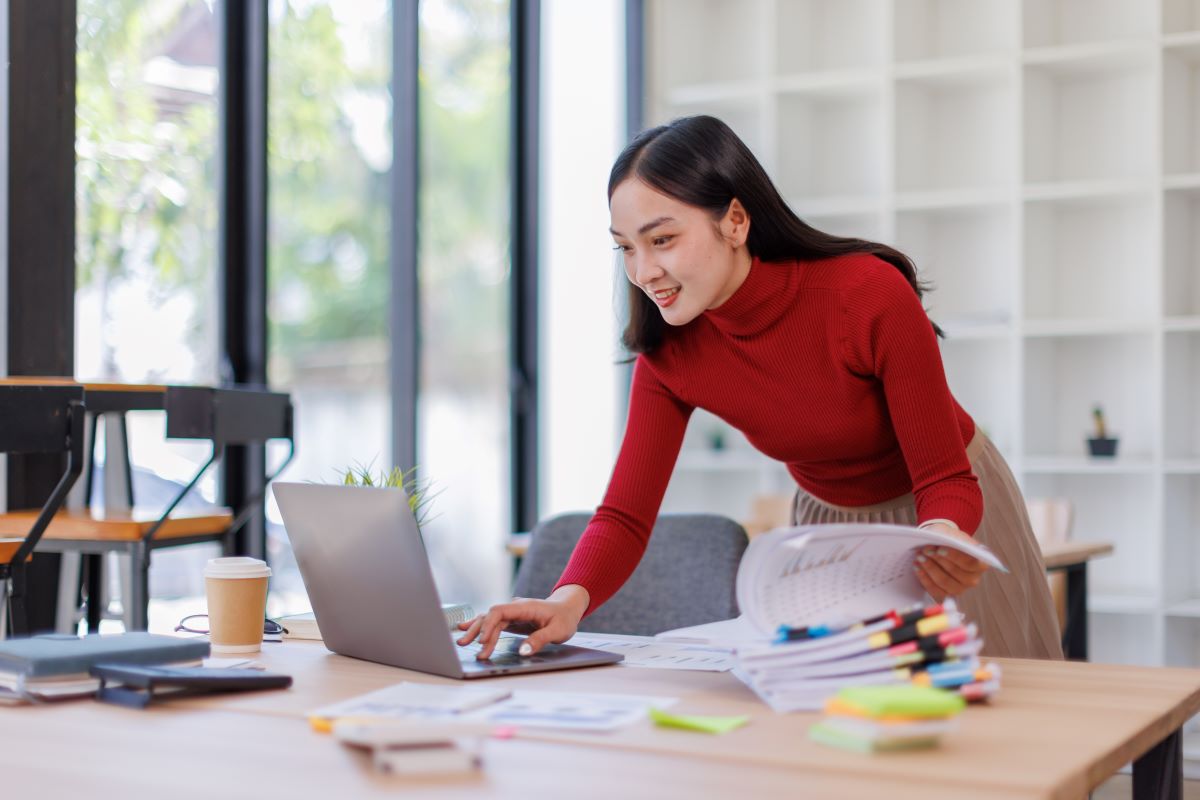 An asian woman works on a laptop with paperwork nearby.